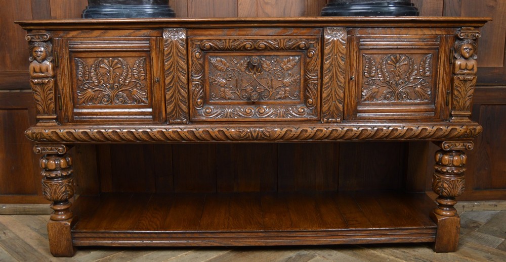 A carved dark oak Edwardian pot board dresser base featuring detailed relief carving on cupboard doors and a wide lower open shelf. View III