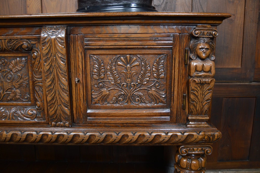 A carved dark oak Edwardian pot board dresser base featuring detailed relief carving on cupboard doors and a wide lower open shelf. View IV