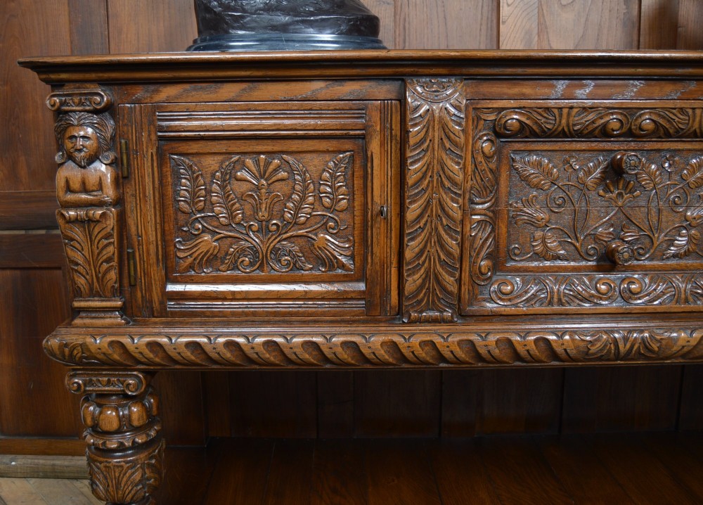 A carved dark oak Edwardian pot board dresser base featuring detailed relief carving on cupboard doors and a wide lower open shelf. View VI