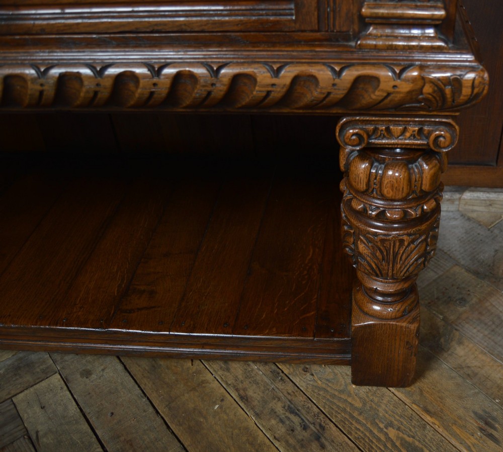 A carved dark oak Edwardian pot board dresser base featuring detailed relief carving on cupboard doors and a wide lower open shelf. View VII