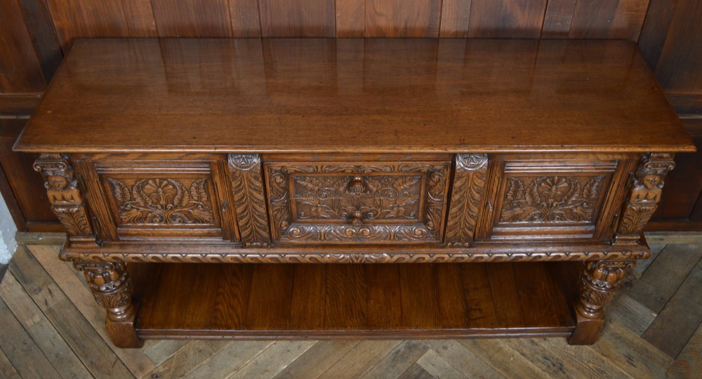 A carved dark oak Edwardian pot board dresser base featuring detailed relief carving on cupboard doors and a wide lower open shelf. View VIII