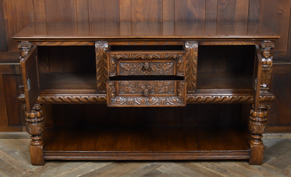 A carved dark oak Edwardian pot board dresser base featuring detailed relief carving on cupboard doors and a wide lower open shelf. View IX