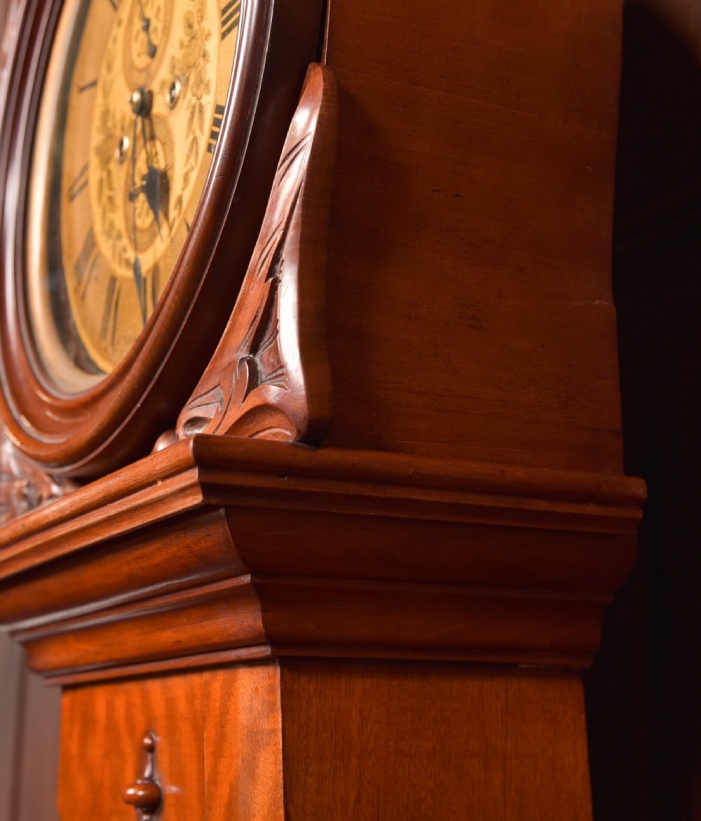 A Victorian Scottish mahogany drumhead longcase clock featuring a circular hood with carved scrollwork, a brass dial, and a flame mahogany trunk. View IX