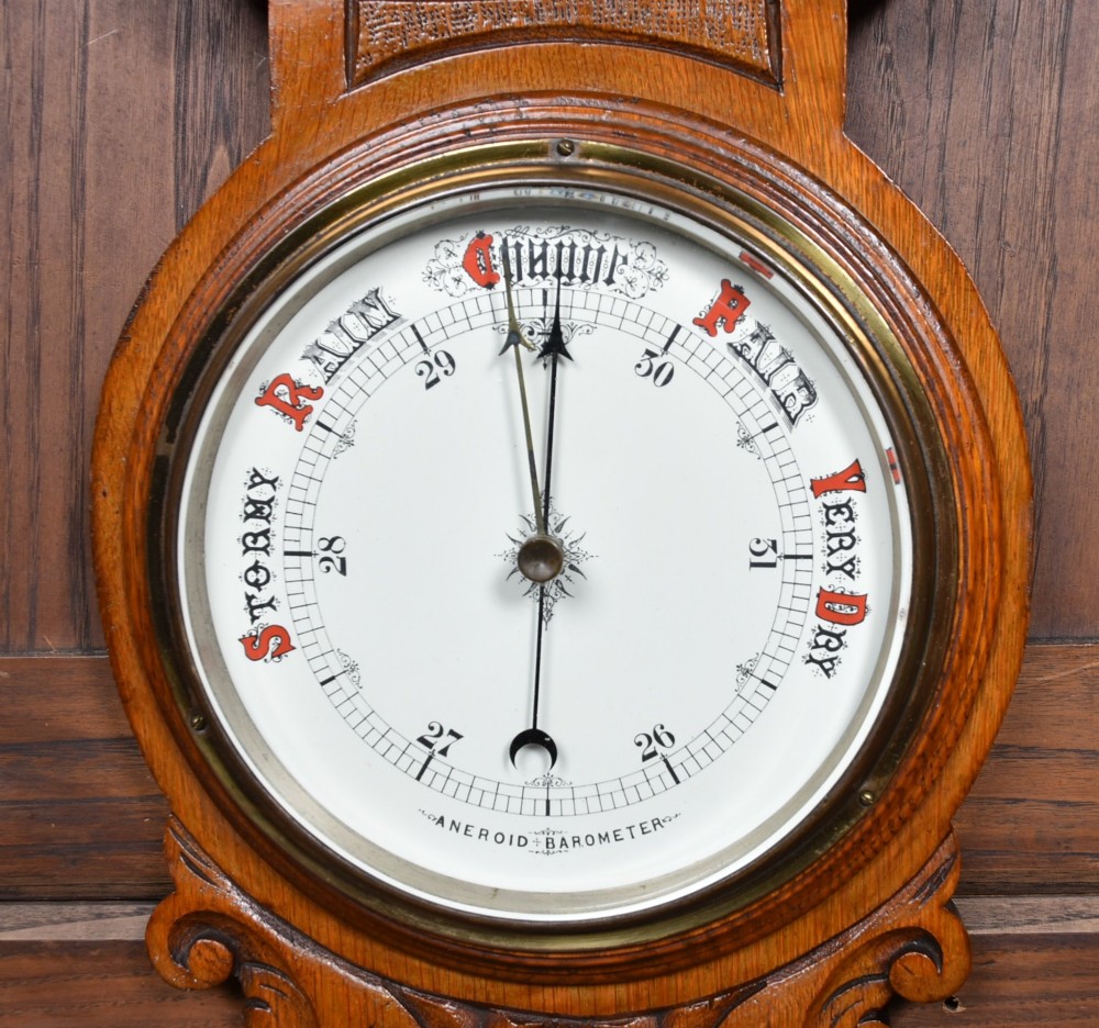 A Victorian carved oak wall barometer featuring an upper thermometer flanked by turned urn finials and a lower round aneroid dial with red and black text. View III