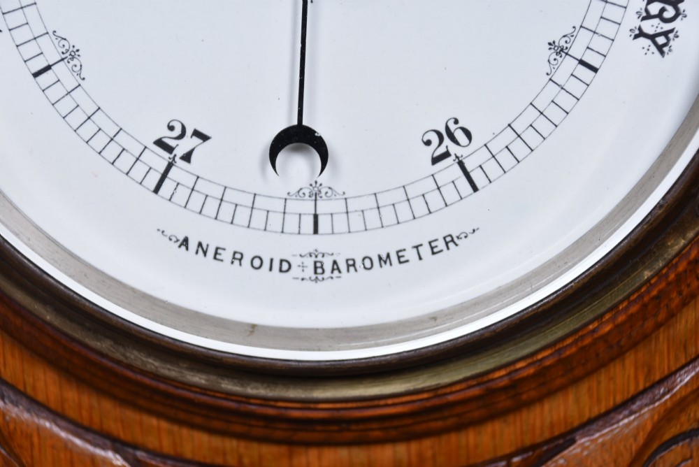 A Victorian carved oak wall barometer featuring an upper thermometer flanked by turned urn finials and a lower round aneroid dial with red and black text. View IV