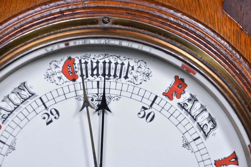 A Victorian carved oak wall barometer featuring an upper thermometer flanked by turned urn finials and a lower round aneroid dial with red and black text. View V