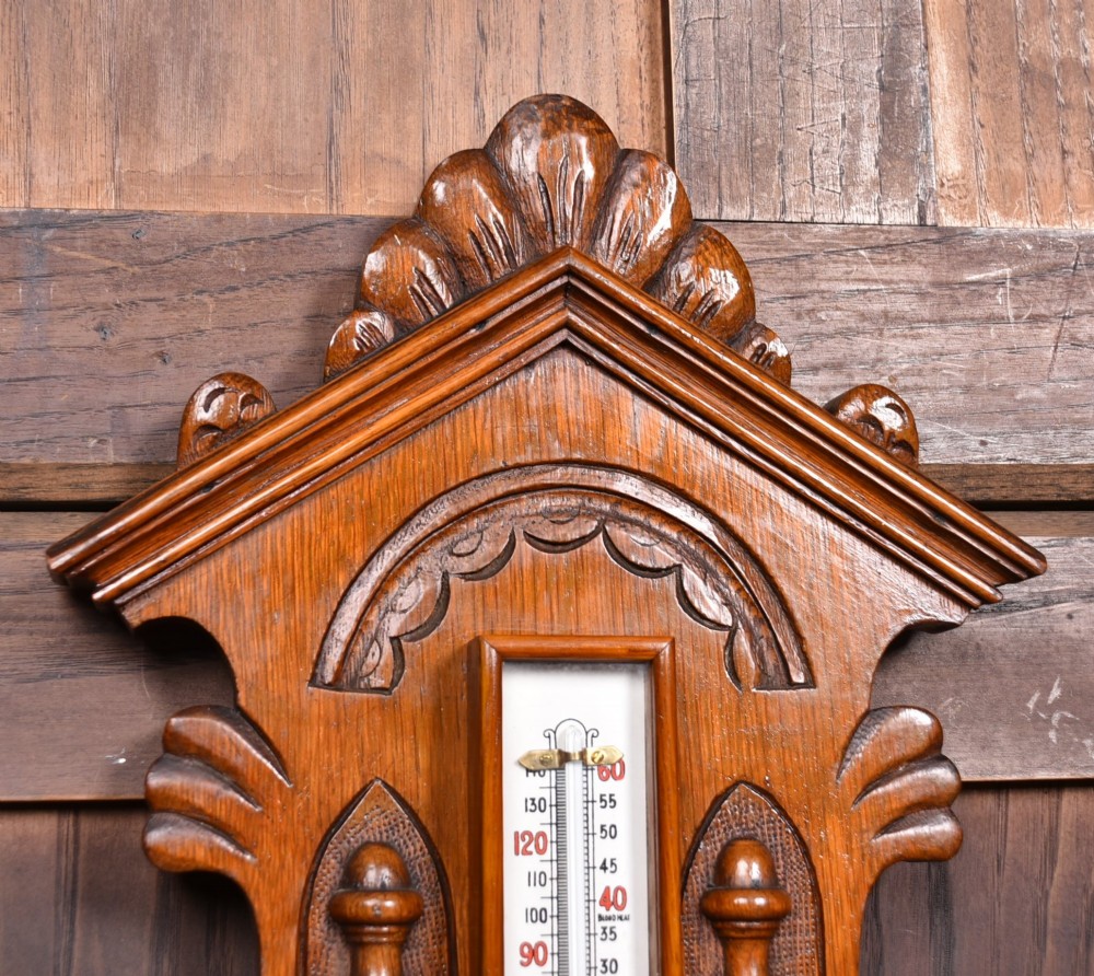 A Victorian carved oak wall barometer featuring an upper thermometer flanked by turned urn finials and a lower round aneroid dial with red and black text. View VII