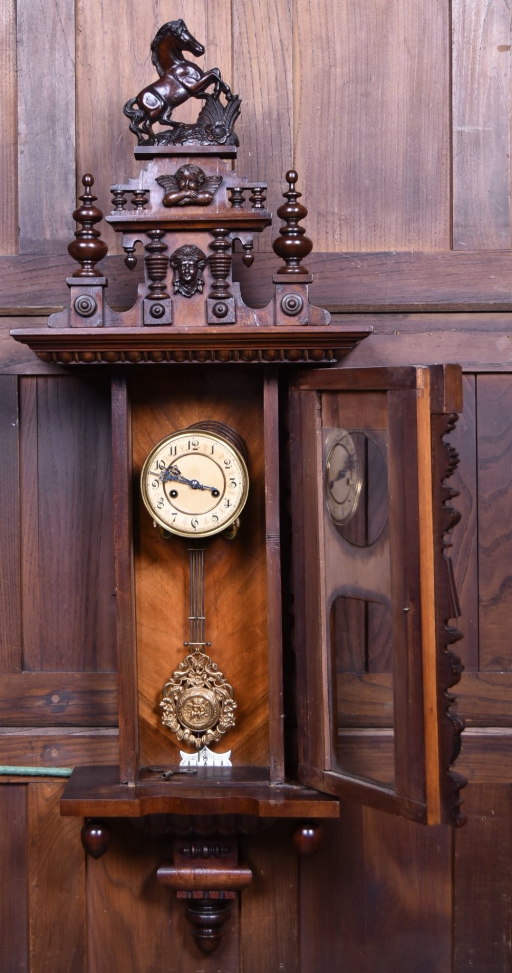 An antique dark walnut wall clock featuring a carved horse crest, turned side pilasters, and a decorative brass pendulum behind a glass door. View IV