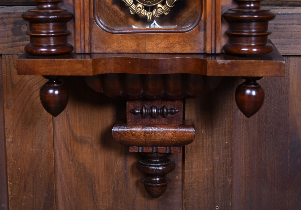 An antique dark walnut wall clock featuring a carved horse crest, turned side pilasters, and a decorative brass pendulum behind a glass door. View VII