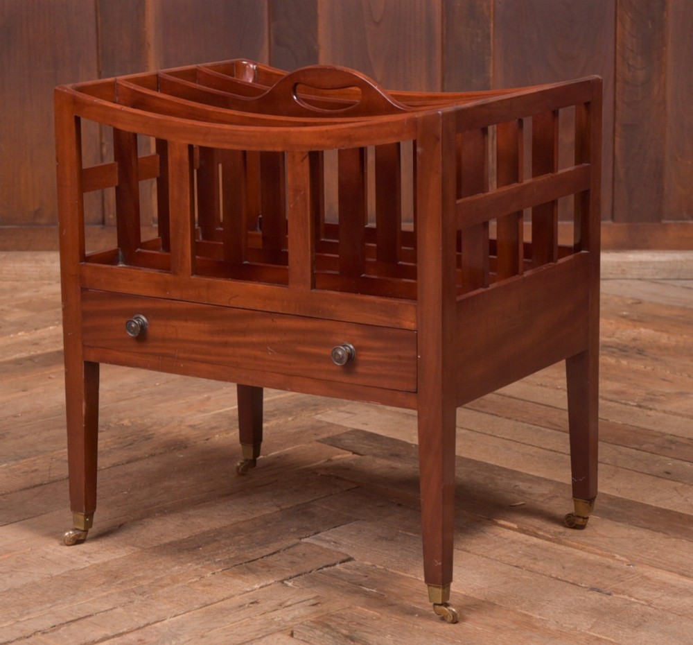 An Edwardian mahogany Canterbury featuring four slatted compartments, an integrated carrying handle, a lower drawer with brass knobs, and square tapered legs on brass castors.