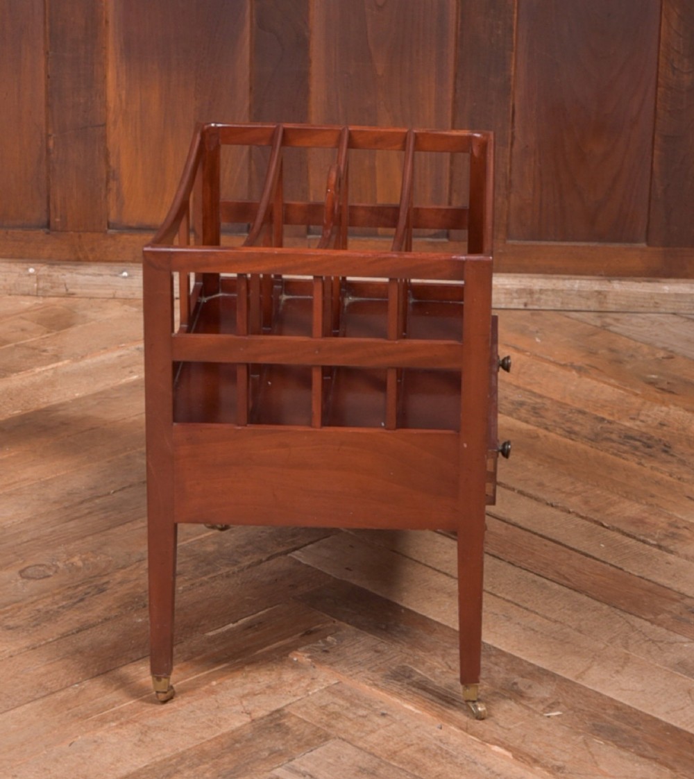 An Edwardian mahogany Canterbury featuring four slatted compartments, an integrated carrying handle, a lower drawer with brass knobs, and square tapered legs on brass castors. View VI