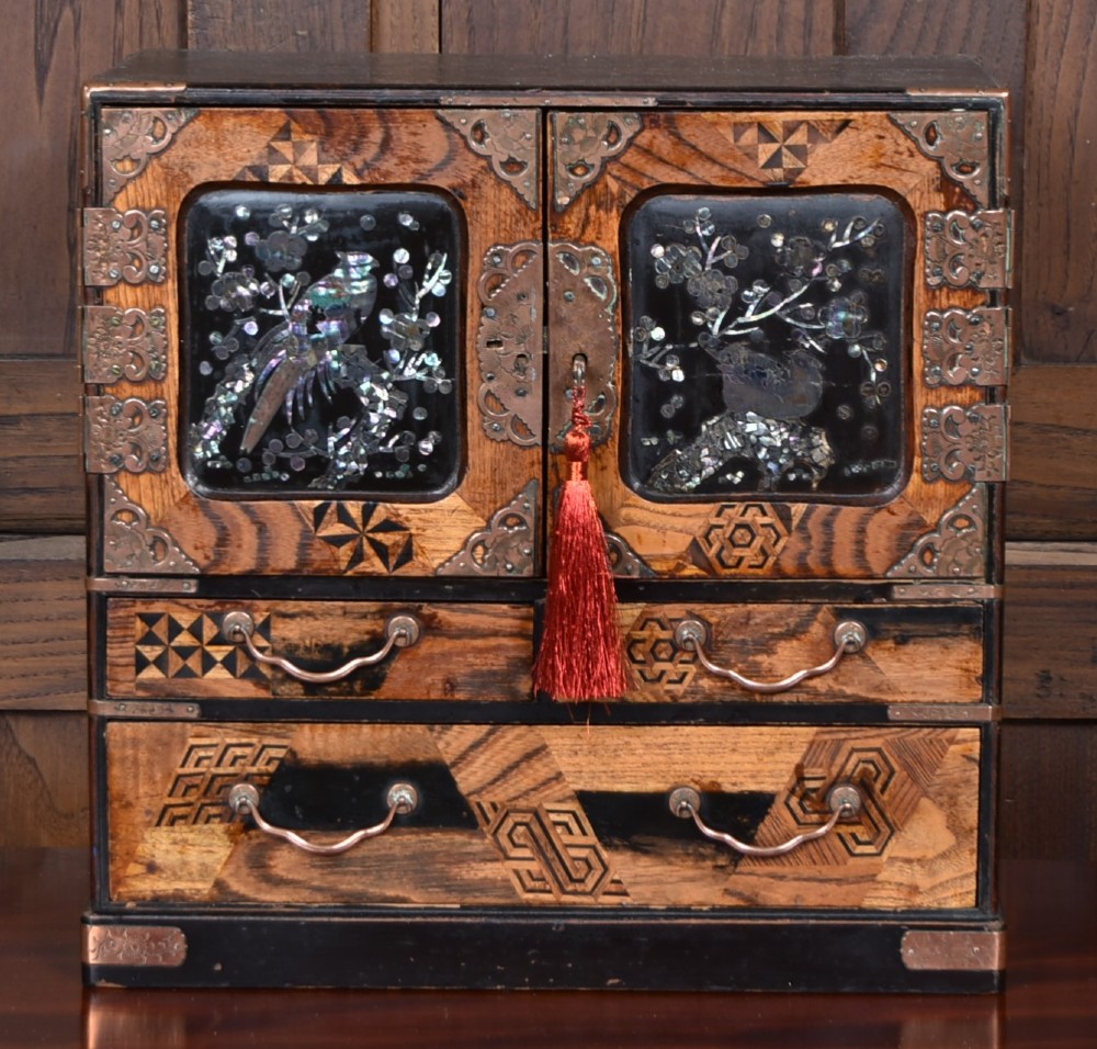 A Japanese tabletop wooden kodansu cabinet featuring geometric marquetry, mother-of-pearl shell inlay on dark lacquered door panels, metal mounts, and lower drawers.