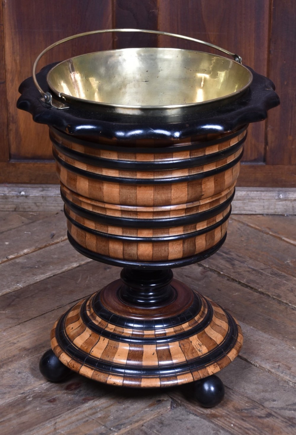 A 19th-century Dutch wooden teestoof featuring alternating light and dark wood parquetry, ebonised trim, turned pedestal base on ball feet, and a removable brass liner with a swing handle.