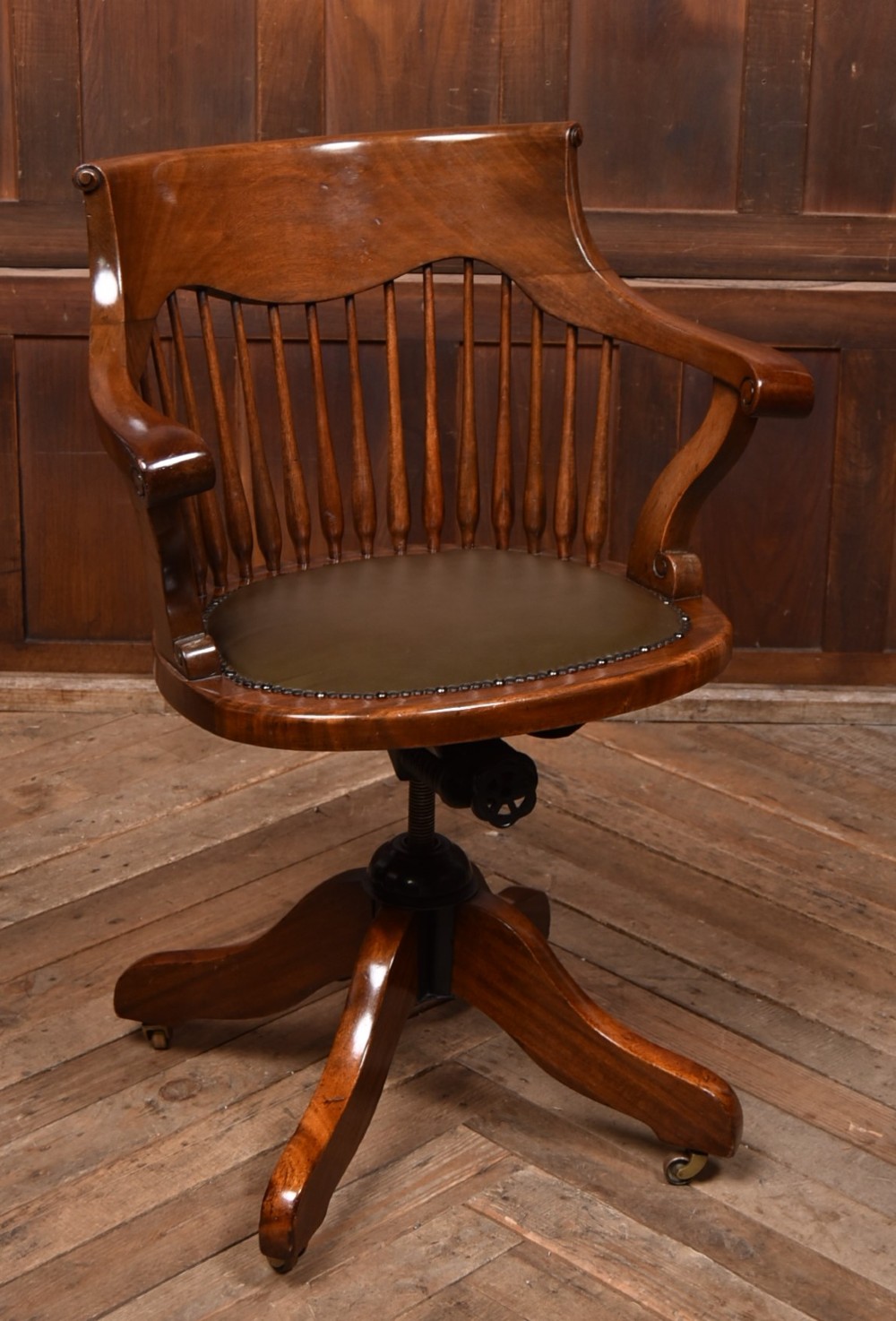 An Edwardian carved mahogany swivel desk chair featuring a spindle backrest, dark leather upholstered seat with studding detail, and a quadruped base on brass castors.
