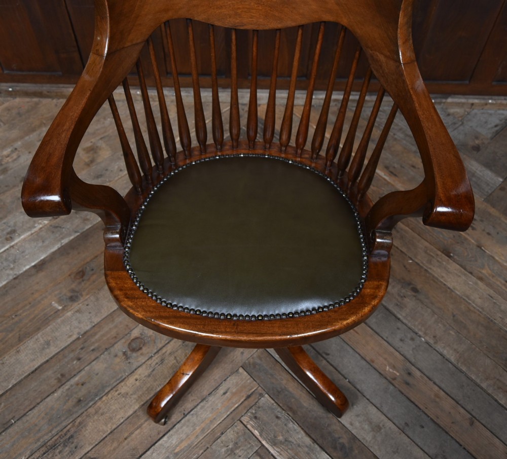An Edwardian carved mahogany swivel desk chair featuring a spindle backrest, dark leather upholstered seat with studding detail, and a quadruped base on brass castors. View V