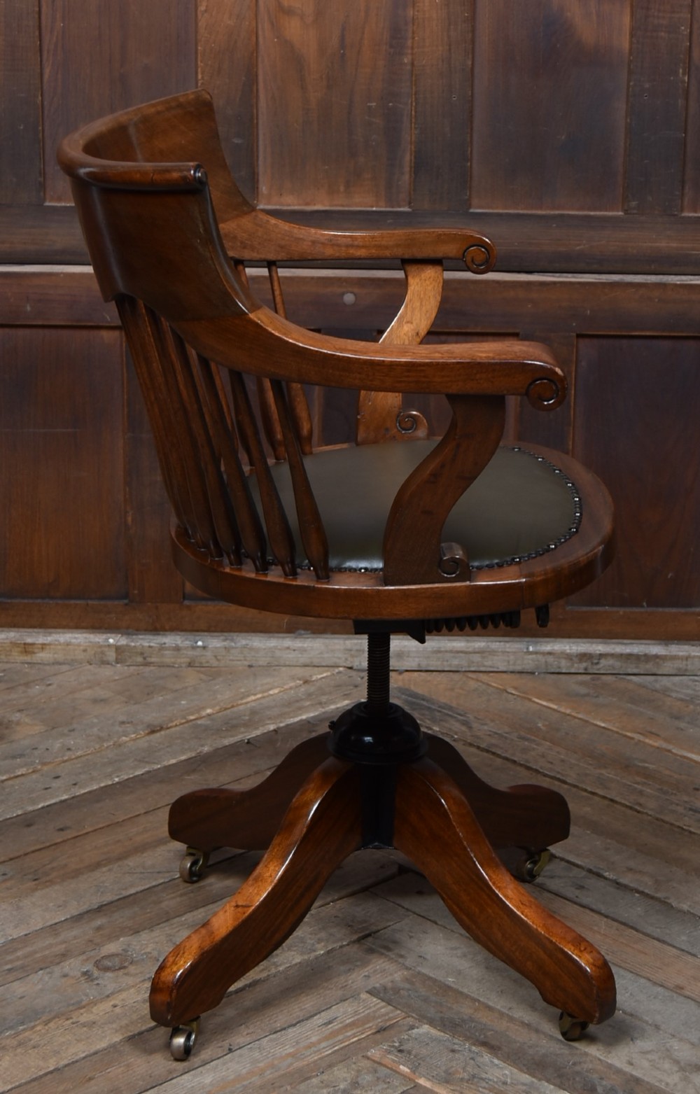 An Edwardian carved mahogany swivel desk chair featuring a spindle backrest, dark leather upholstered seat with studding detail, and a quadruped base on brass castors. View VI
