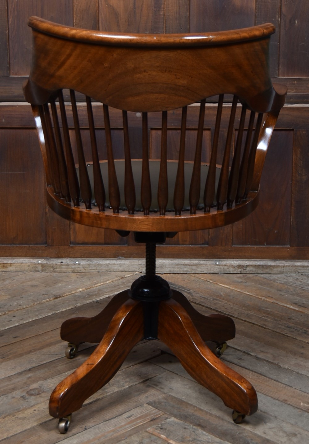 An Edwardian carved mahogany swivel desk chair featuring a spindle backrest, dark leather upholstered seat with studding detail, and a quadruped base on brass castors. View VII