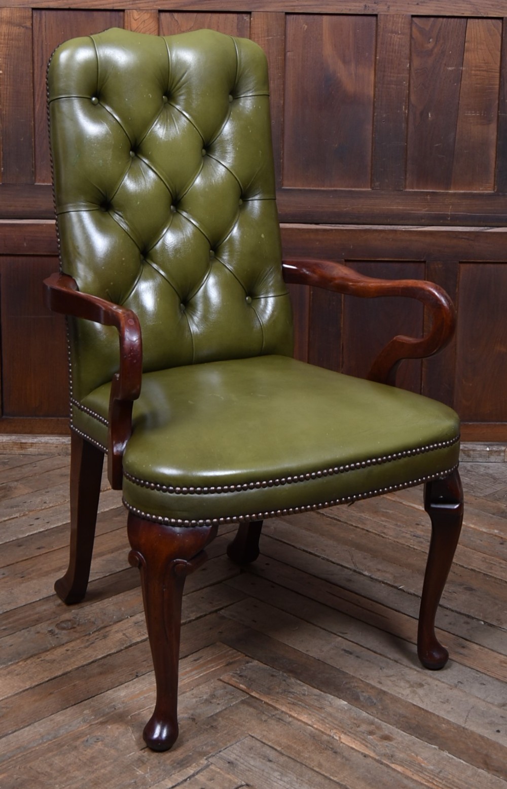A high-backed desk chair featuring olive green button-tufted leather upholstery, carved dark wood armrests, brass stud trimming, and front cabriole legs.