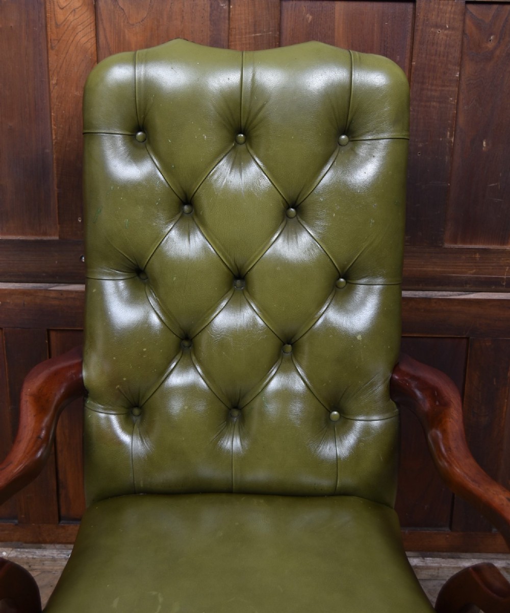 A high-backed desk chair featuring olive green button-tufted leather upholstery, carved dark wood armrests, brass stud trimming, and front cabriole legs. View VI