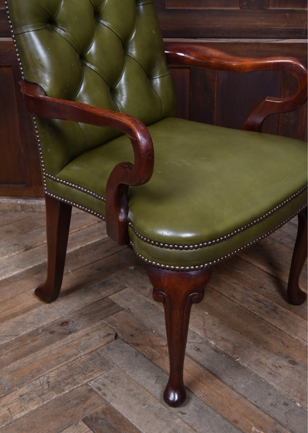 A high-backed desk chair featuring olive green button-tufted leather upholstery, carved dark wood armrests, brass stud trimming, and front cabriole legs. View VII