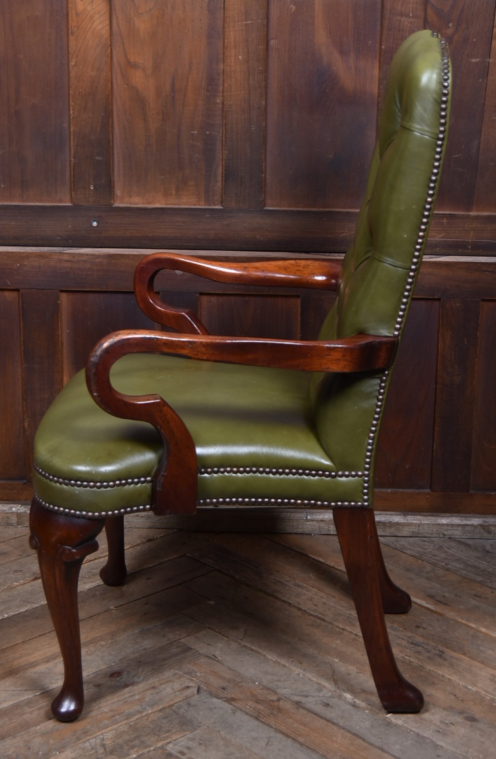 A high-backed desk chair featuring olive green button-tufted leather upholstery, carved dark wood armrests, brass stud trimming, and front cabriole legs. View X