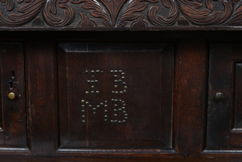 A 17th-century dark oak court cupboard featuring a carved lunette frieze, turned drop pendants, studded panel initials, and geometric lower doors. View II