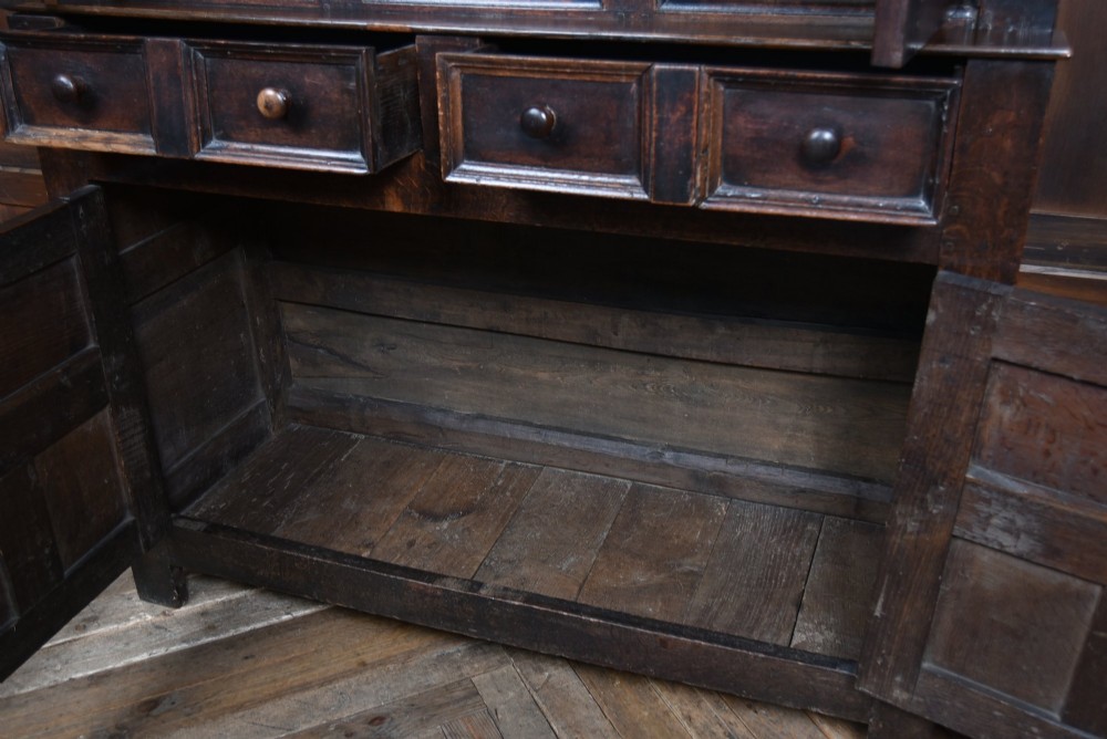 A 17th-century dark oak court cupboard featuring a carved lunette frieze, turned drop pendants, studded panel initials, and geometric lower doors. View IV