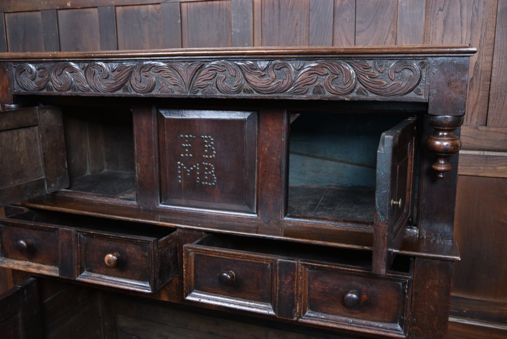 A 17th-century dark oak court cupboard featuring a carved lunette frieze, turned drop pendants, studded panel initials, and geometric lower doors. View V