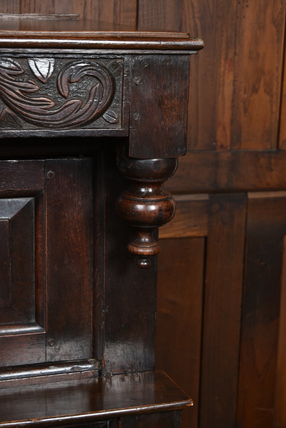 A 17th-century dark oak court cupboard featuring a carved lunette frieze, turned drop pendants, studded panel initials, and geometric lower doors. View VII