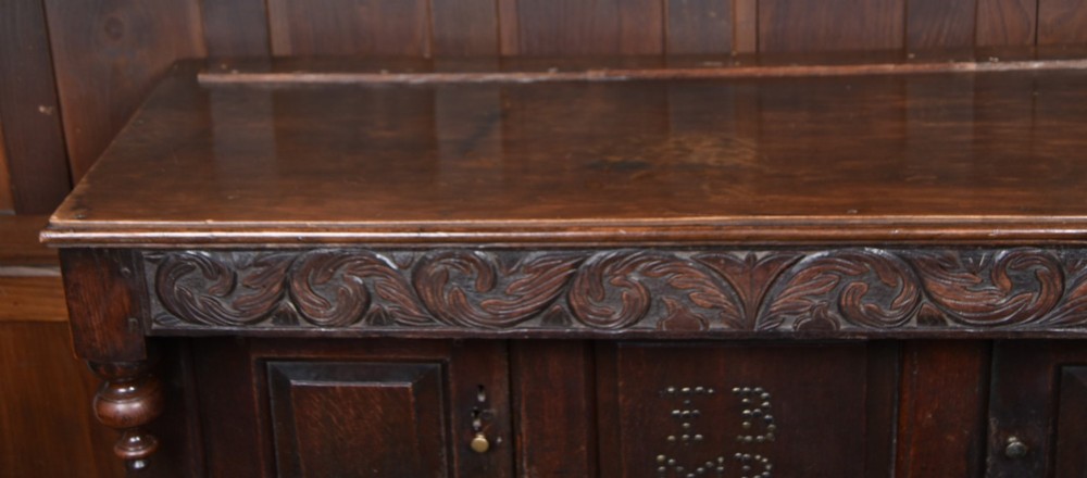 A 17th-century dark oak court cupboard featuring a carved lunette frieze, turned drop pendants, studded panel initials, and geometric lower doors. View XIII