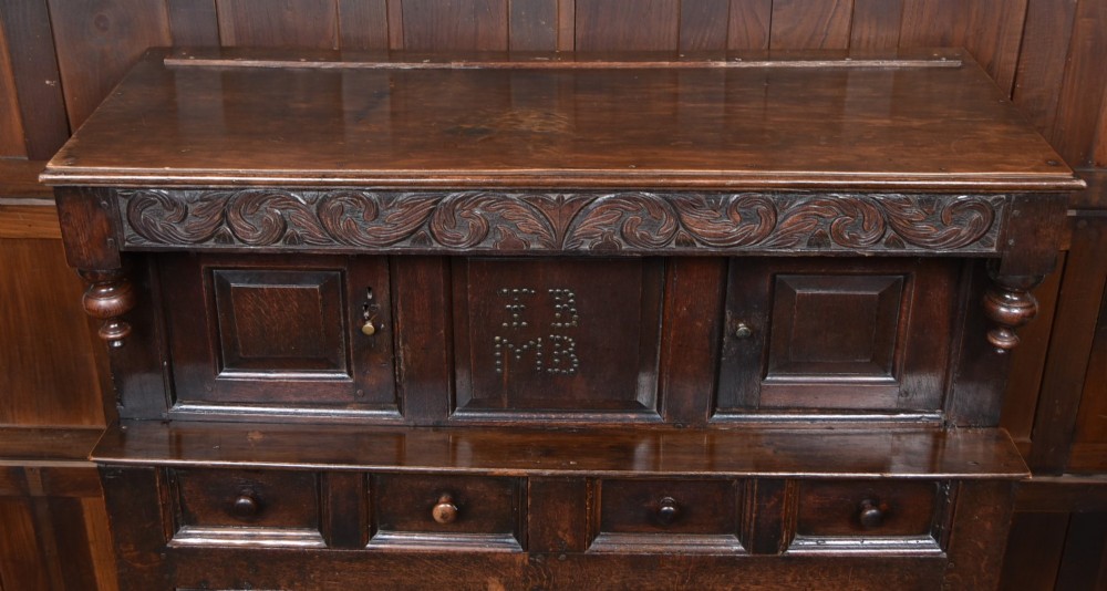 A 17th-century dark oak court cupboard featuring a carved lunette frieze, turned drop pendants, studded panel initials, and geometric lower doors. View XIV