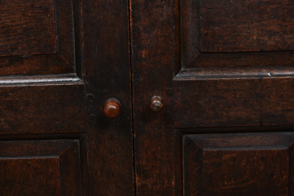A 17th-century dark oak court cupboard featuring a carved lunette frieze, turned drop pendants, studded panel initials, and geometric lower doors. View XV