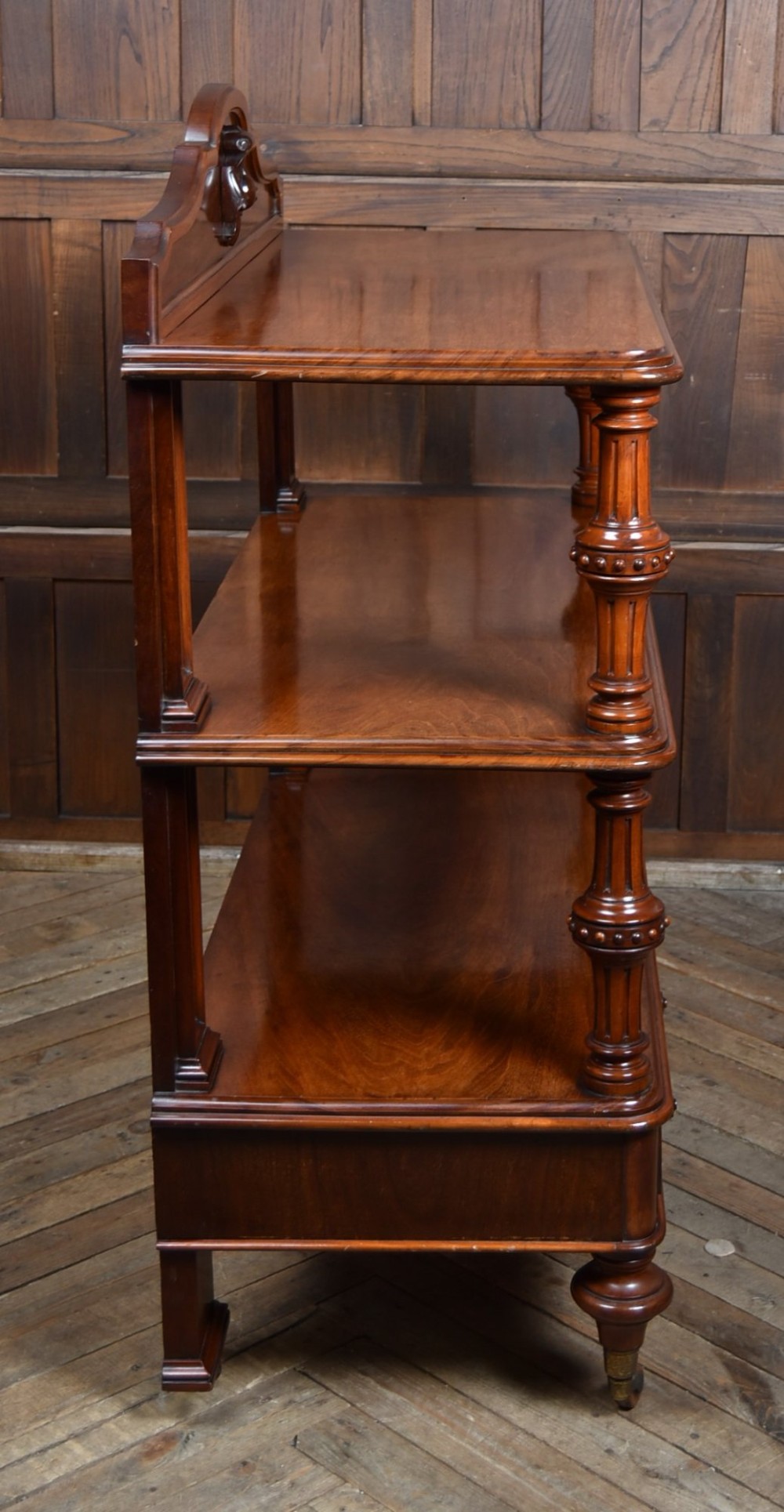 A Victorian mahogany three-tiered whatnot featuring turned column supports, two lower drawers with knob handles, an arched gallery top, and brass castors. View XI