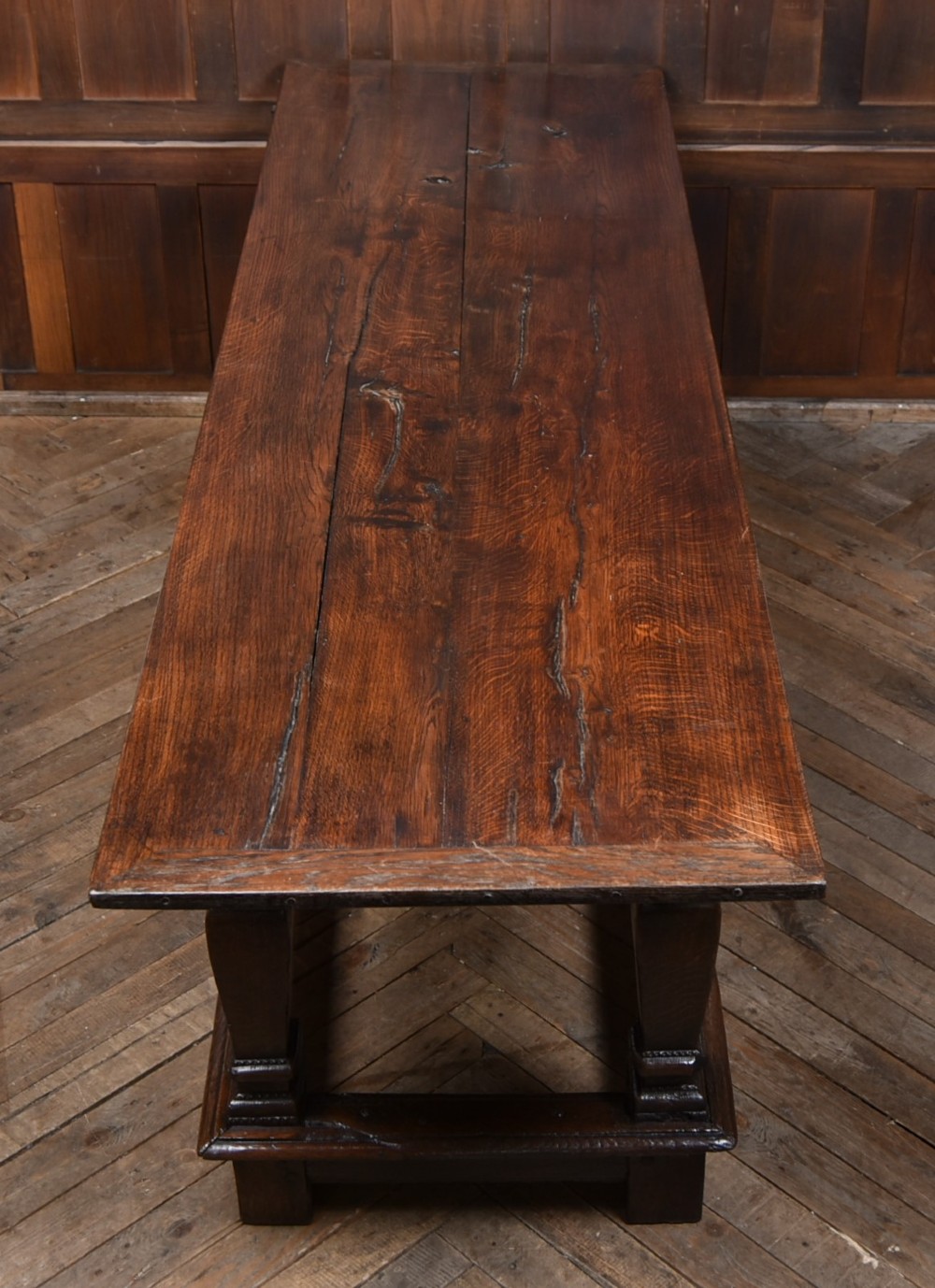 An 18th-century dark oak refectory dining table featuring a long plank top, scalloped apron carving, turned legs, and low foot stretchers. View III