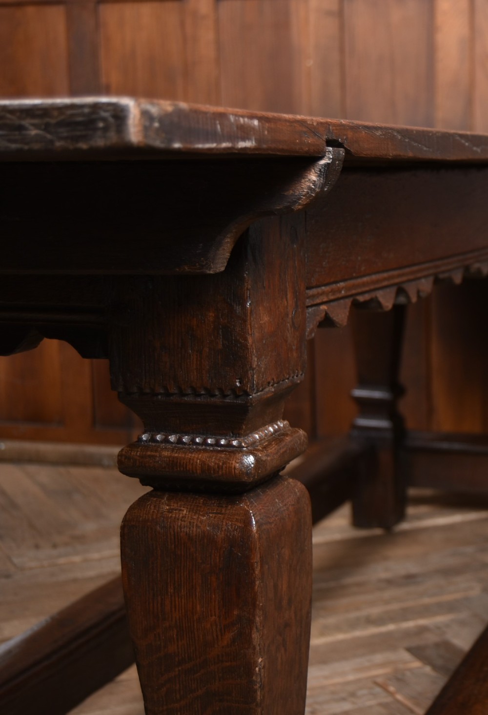 An 18th-century dark oak refectory dining table featuring a long plank top, scalloped apron carving, turned legs, and low foot stretchers. View V
