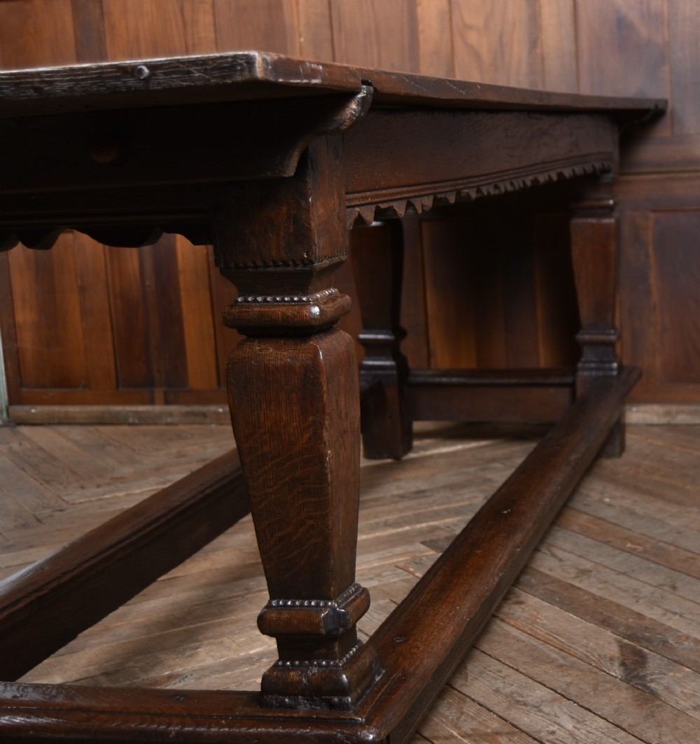 An 18th-century dark oak refectory dining table featuring a long plank top, scalloped apron carving, turned legs, and low foot stretchers. View VI