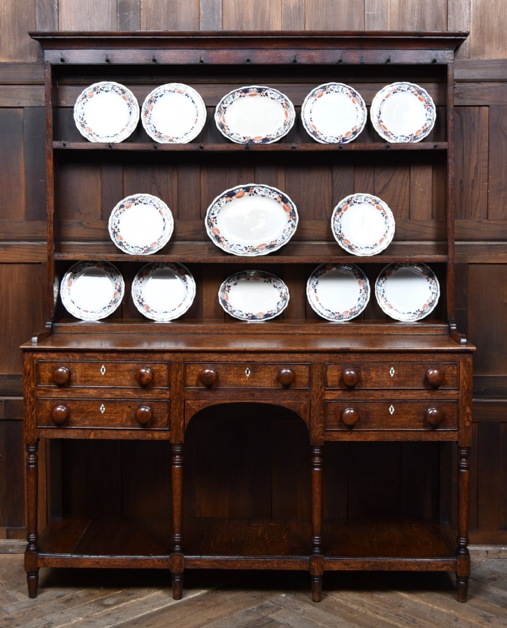 A dark patinated oak country dresser featuring a three-tiered open plate rack stacked with ceramic tableware, set above a base with five drawers, turned front legs, and a low pot board shelf.