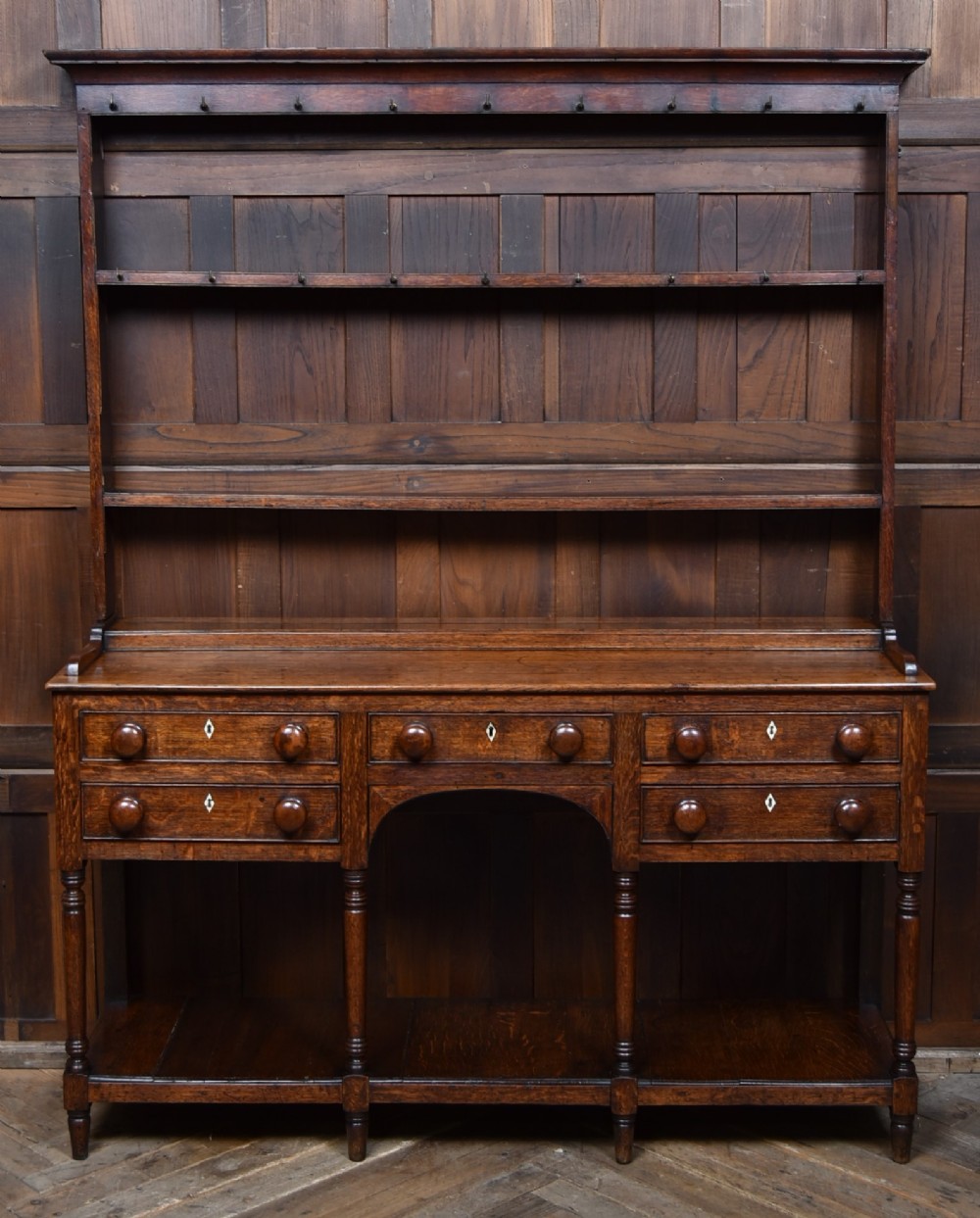 A dark patinated oak country dresser featuring a three-tiered open plate rack stacked with ceramic tableware, set above a base with five drawers, turned front legs, and a low pot board shelf. View II