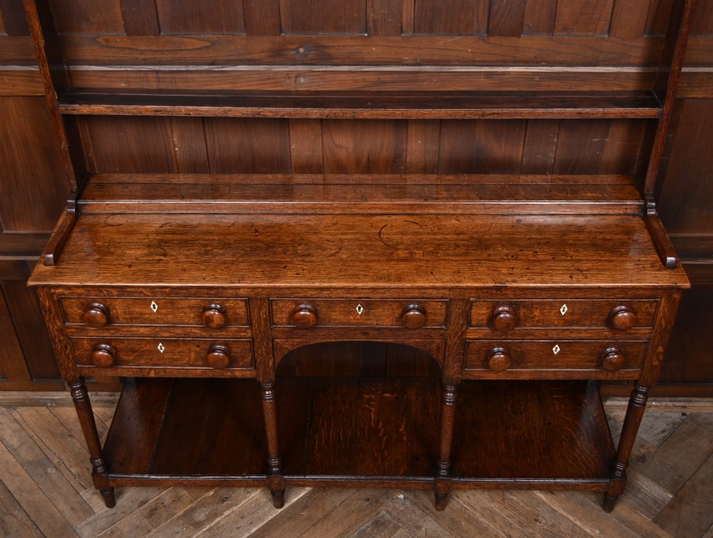 A dark patinated oak country dresser featuring a three-tiered open plate rack stacked with ceramic tableware, set above a base with five drawers, turned front legs, and a low pot board shelf. View IV