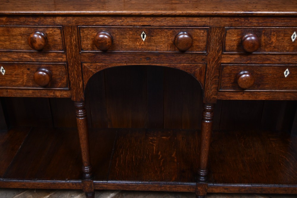 A dark patinated oak country dresser featuring a three-tiered open plate rack stacked with ceramic tableware, set above a base with five drawers, turned front legs, and a low pot board shelf. View VI