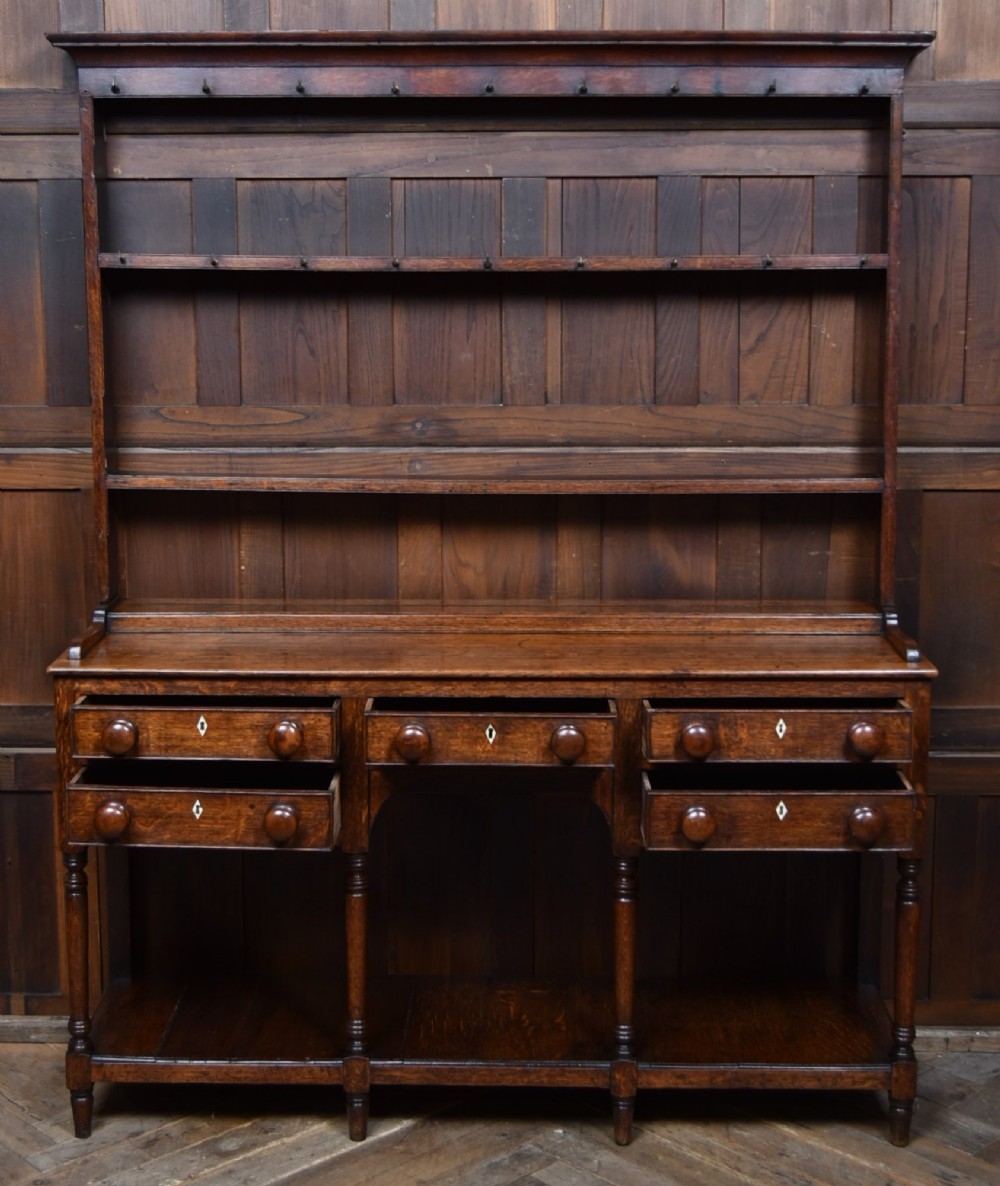 A dark patinated oak country dresser featuring a three-tiered open plate rack stacked with ceramic tableware, set above a base with five drawers, turned front legs, and a low pot board shelf. View VII