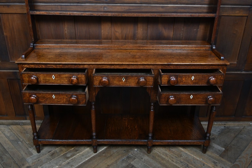 A dark patinated oak country dresser featuring a three-tiered open plate rack stacked with ceramic tableware, set above a base with five drawers, turned front legs, and a low pot board shelf. View VIII