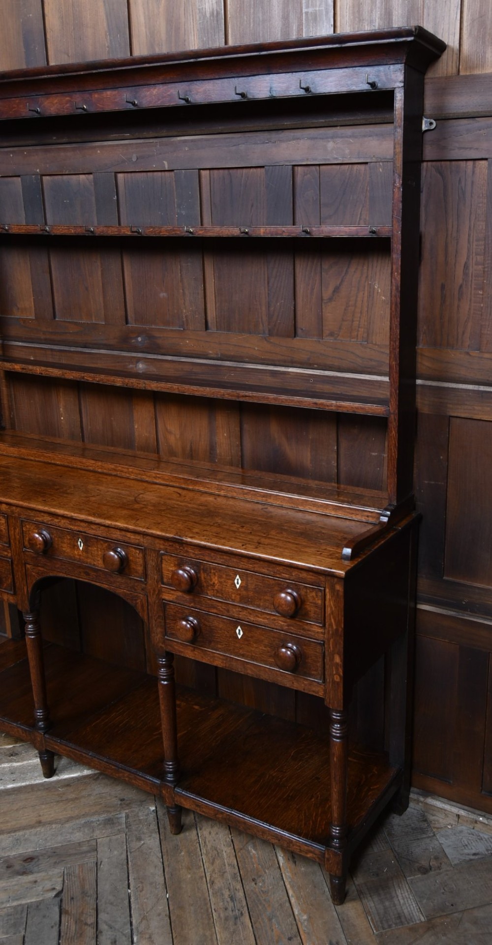 A dark patinated oak country dresser featuring a three-tiered open plate rack stacked with ceramic tableware, set above a base with five drawers, turned front legs, and a low pot board shelf. View IX