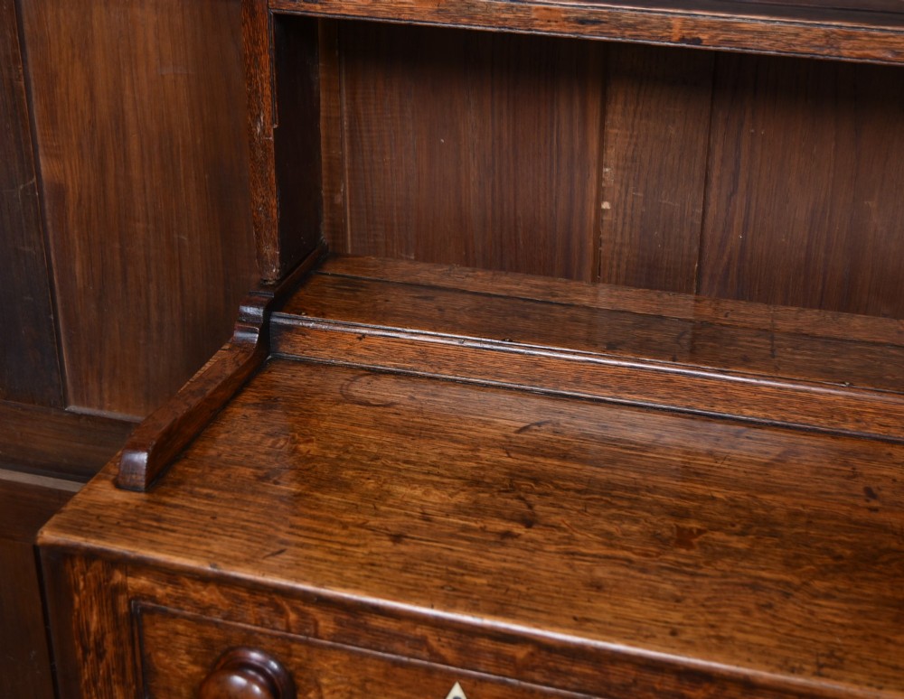 A dark patinated oak country dresser featuring a three-tiered open plate rack stacked with ceramic tableware, set above a base with five drawers, turned front legs, and a low pot board shelf. View XI