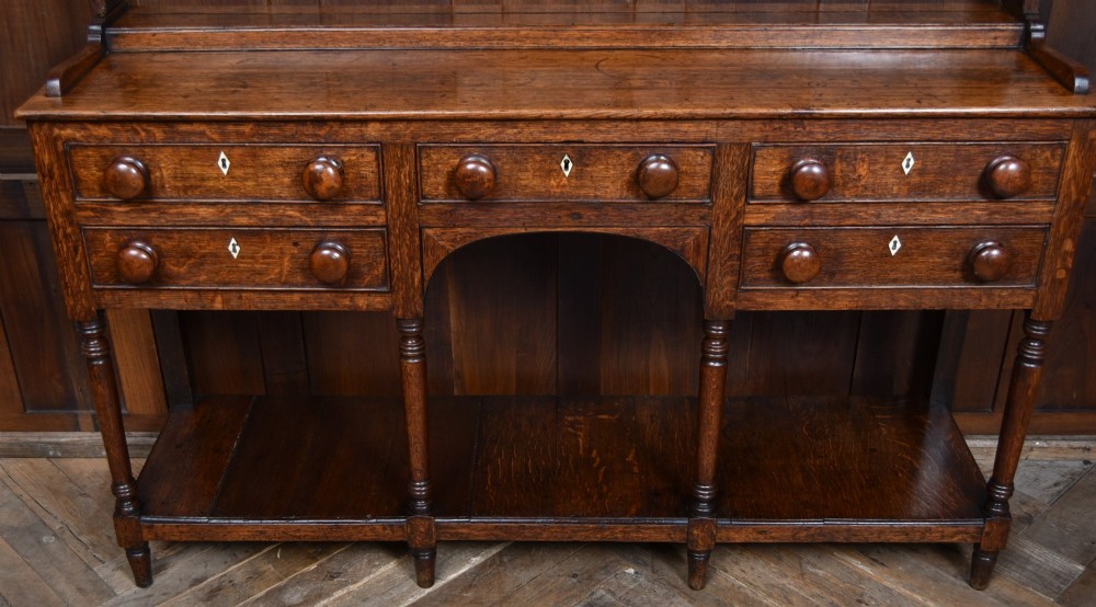 A dark patinated oak country dresser featuring a three-tiered open plate rack stacked with ceramic tableware, set above a base with five drawers, turned front legs, and a low pot board shelf. View XII