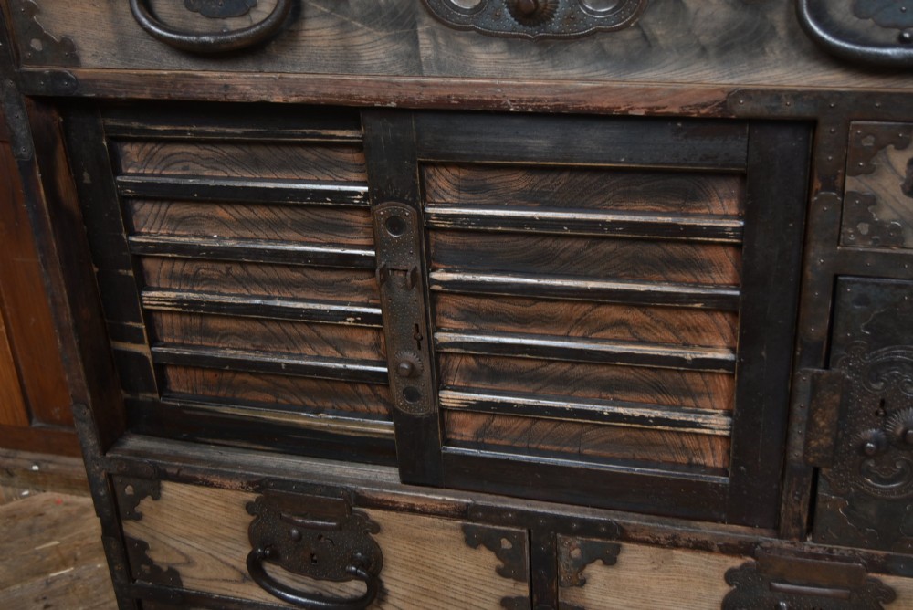 A Japanese tansu wooden chest featuring multiple drawers, sliding louvered doors, a small lockable door, and dark iron corner plates and drop handles. View V