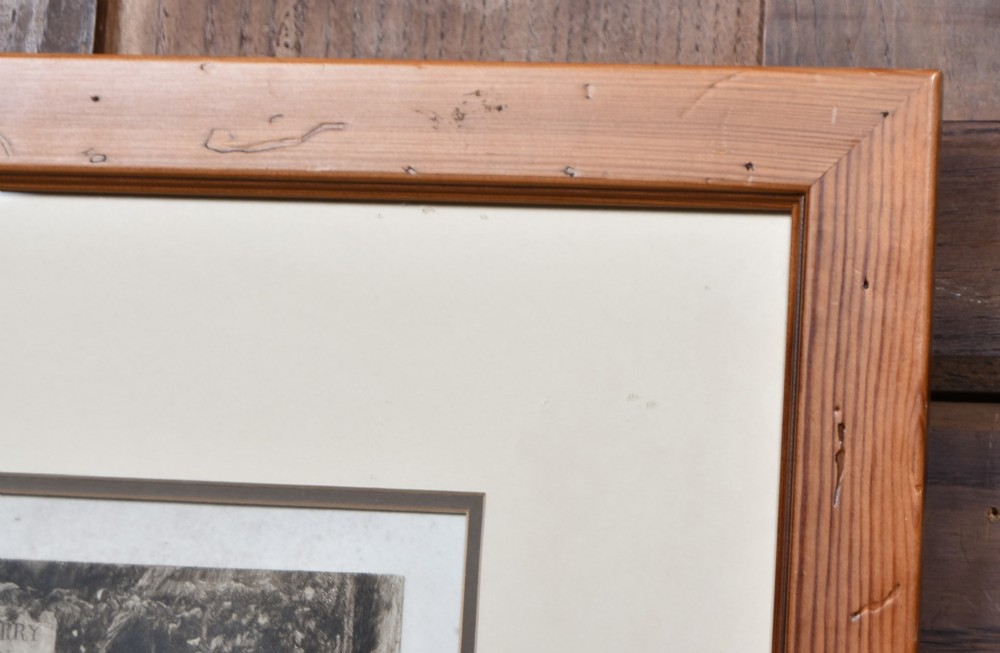 A framed 19th-century monochrome etching depicting figures, a horse, and a dog beside a river outside a village inn, encased in a light wood frame with an off-white mount. View X