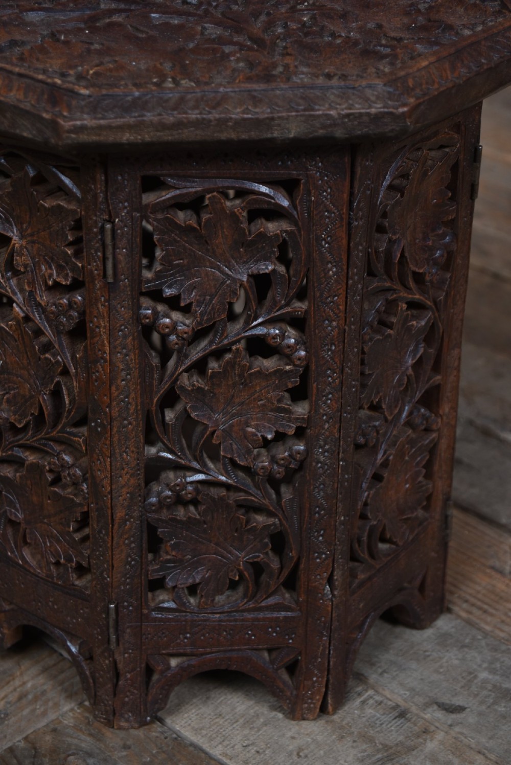 An octagonal dark hardwood side table featuring deep relief foliate carving across the top and openwork pierced panels on the folding base. View II
