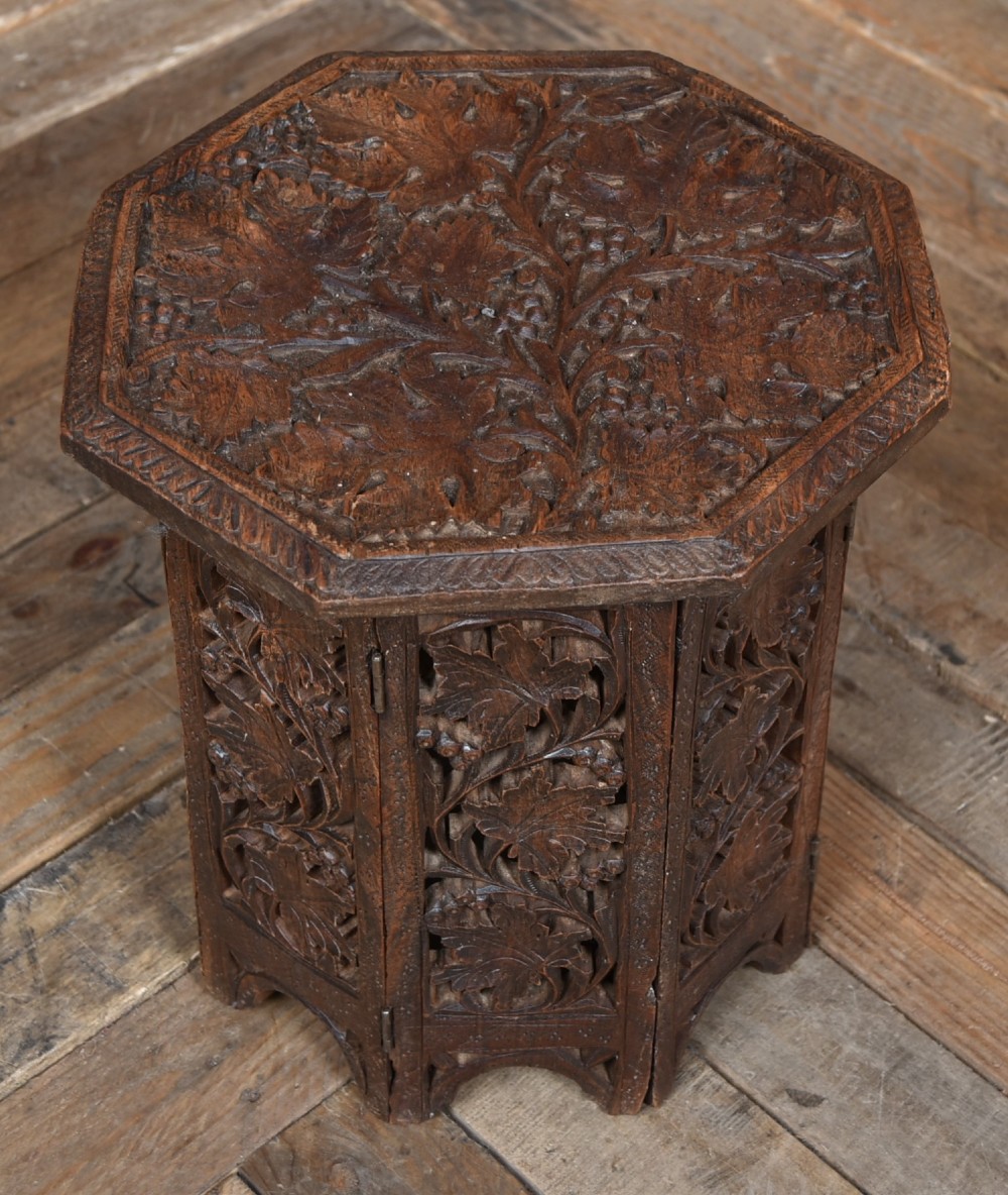 An octagonal dark hardwood side table featuring deep relief foliate carving across the top and openwork pierced panels on the folding base. View III