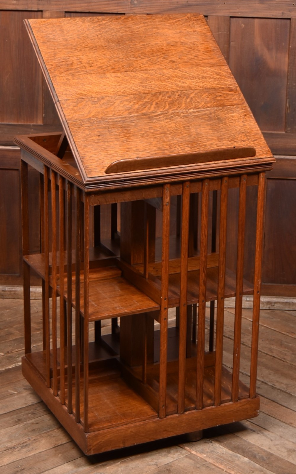 A two-tier freestanding timber revolving bookcase with slatted sides and an angled reading lectern top holding an open volume. View II